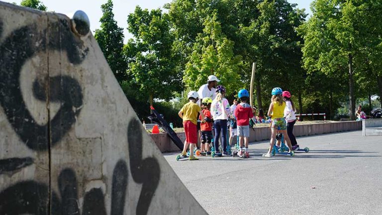 Kinder stehen mit ihren Rollern im Niddapark in Frankfurt im Skatepark und lernen beim Fun Wheel Camp der Outdoorkids, wie man mit dem Roller umgeht.