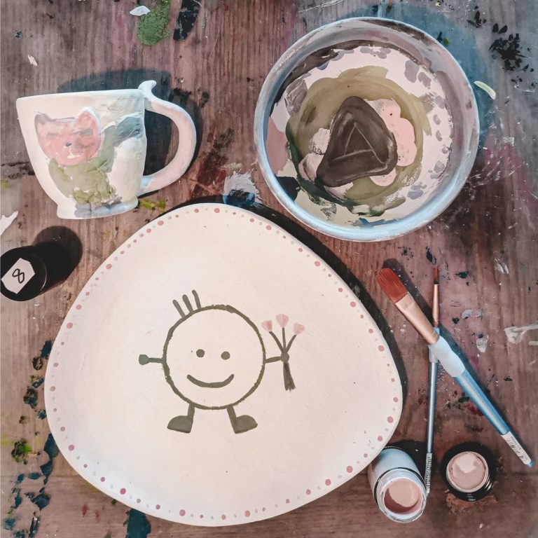 Coloria Frankfurt - eine schön bemalte Tasse, eine Müslischale und ein Teller liegen fertig bemalt auf dem Tisch. Überall herum liegen Farben und Pinsel, um die Keramik selbst zu bemalen.