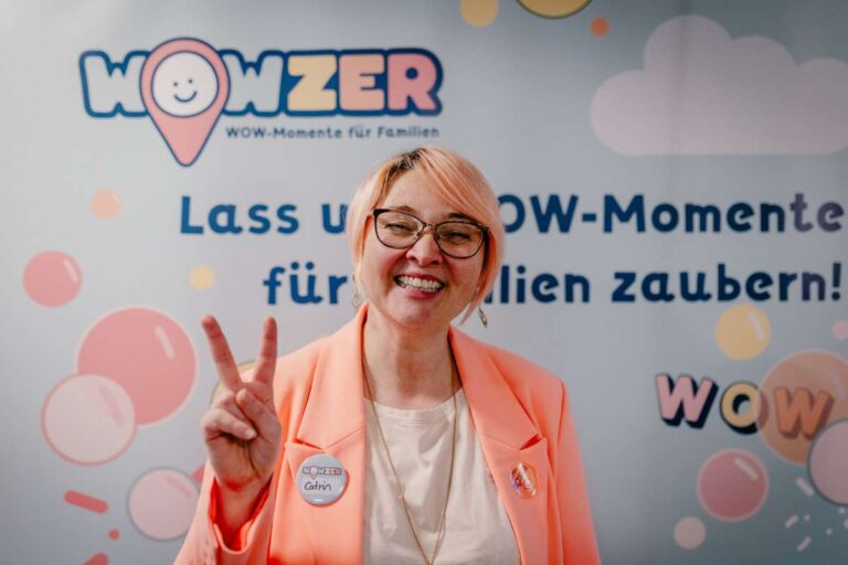 Catrin von Frankfurt mit Kids steht mit einem Peace Zeichen vor der Leinwand mit WOWZER, auf der steht: Lass uns WOW-Momente für Familien zaubern